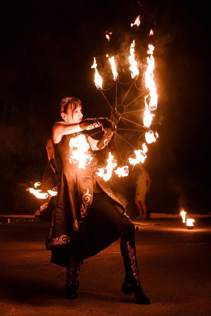 Performer dressed in ornate outfit spinning a wheel with multiple flaming torches at night.